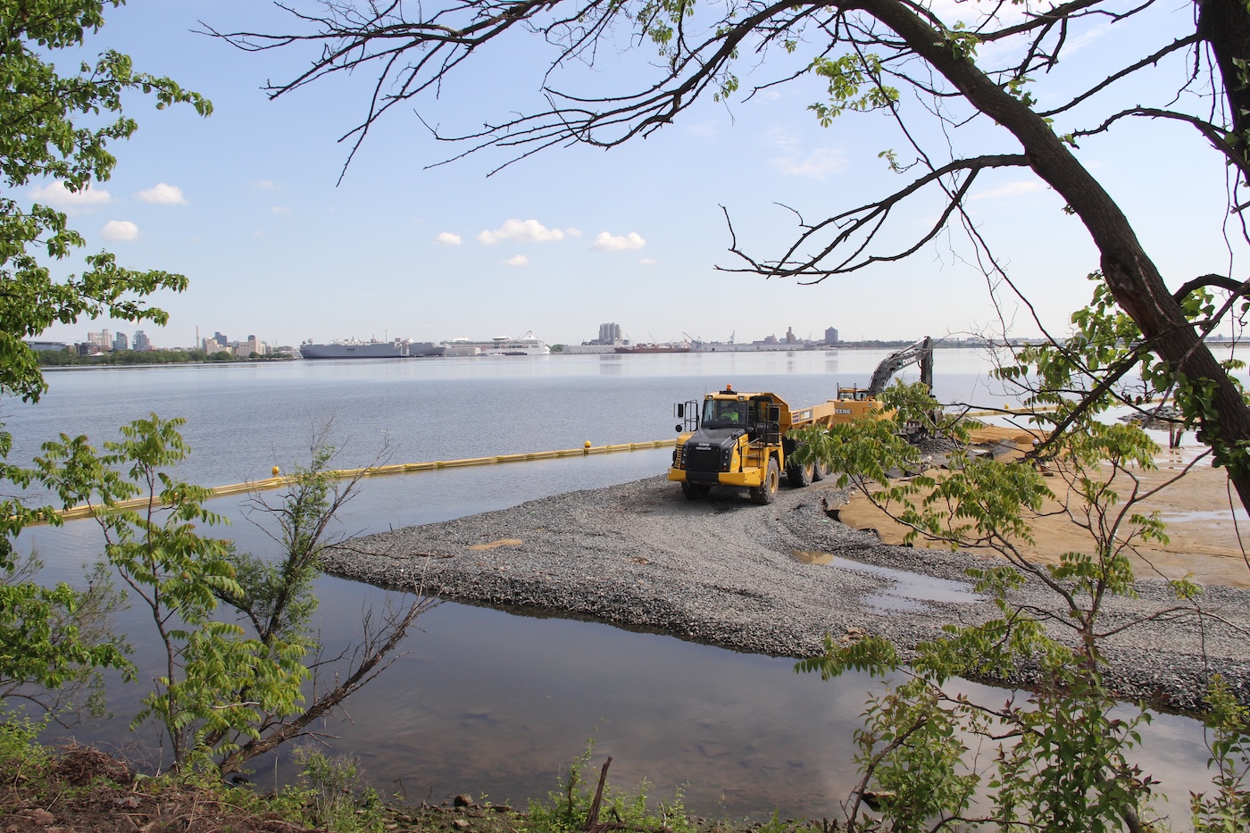 Photo of shoreline, with construction vehicle placing stone and sand in water, and trees in foreground.